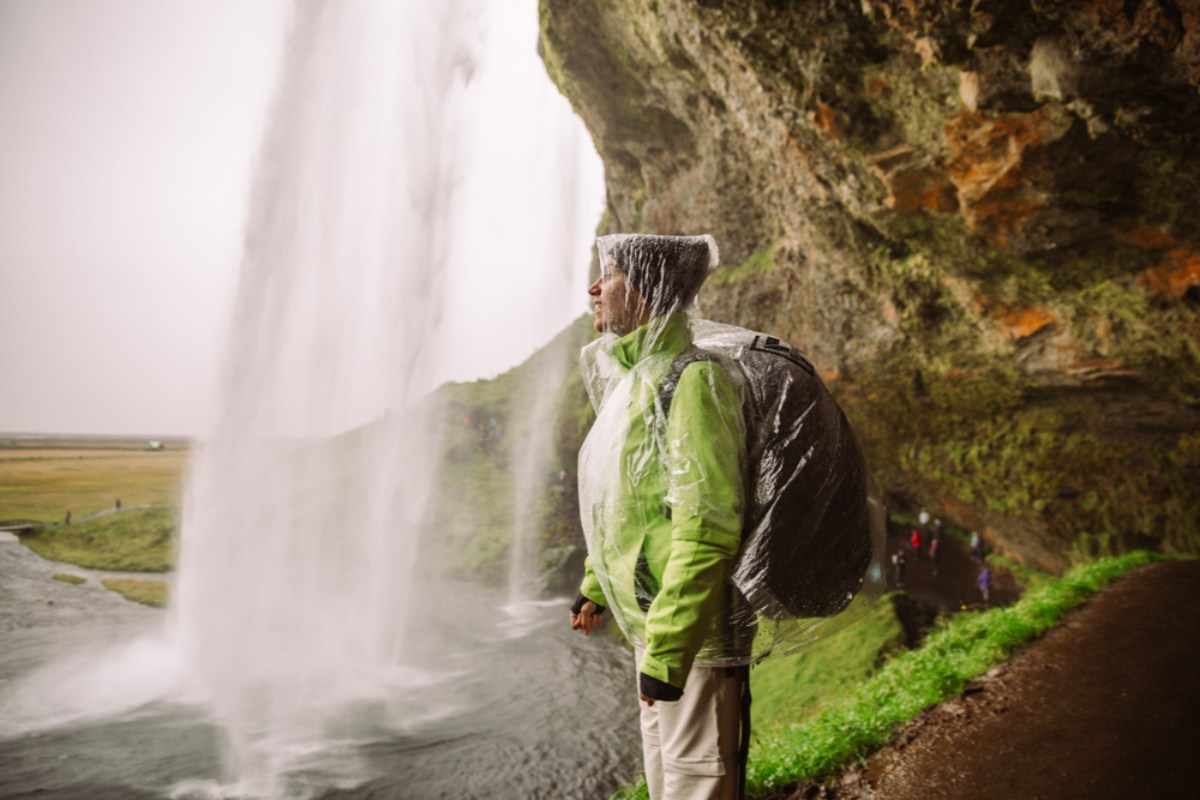 Traveler in a rain shell and poncho near a waterfall, mist swirling all around.