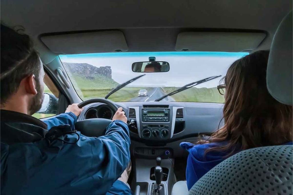 Two people inside a rental car driving on a wet road in Iceland, windshield wipers on, with green fields and rocky hills visible through the windshiel