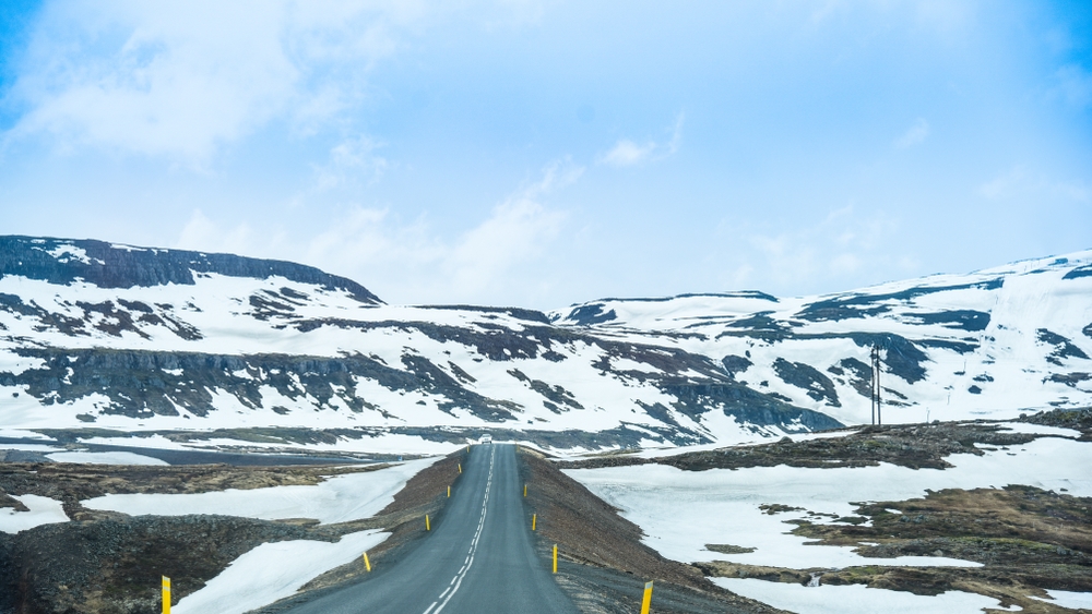 Two-lane Icelandic mountain road with yellow markers cutting through snow-covered hills under a blue sky.