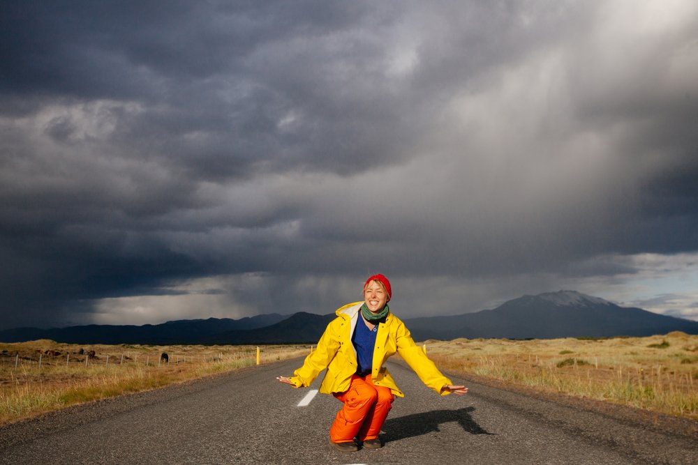 Traveler in yellow jacket kneeling on an empty Icelandic road under dramatic storm clouds.