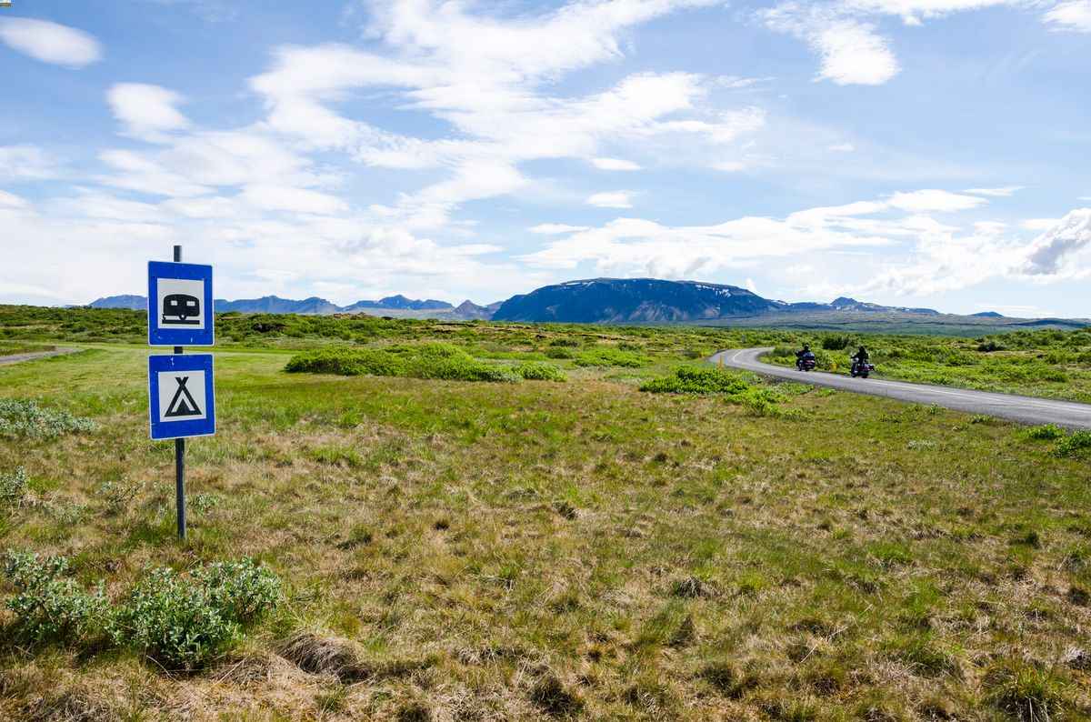 Roadside blue camping and motorhome signs on an open Icelandic landscape with a winding road and distant riders.