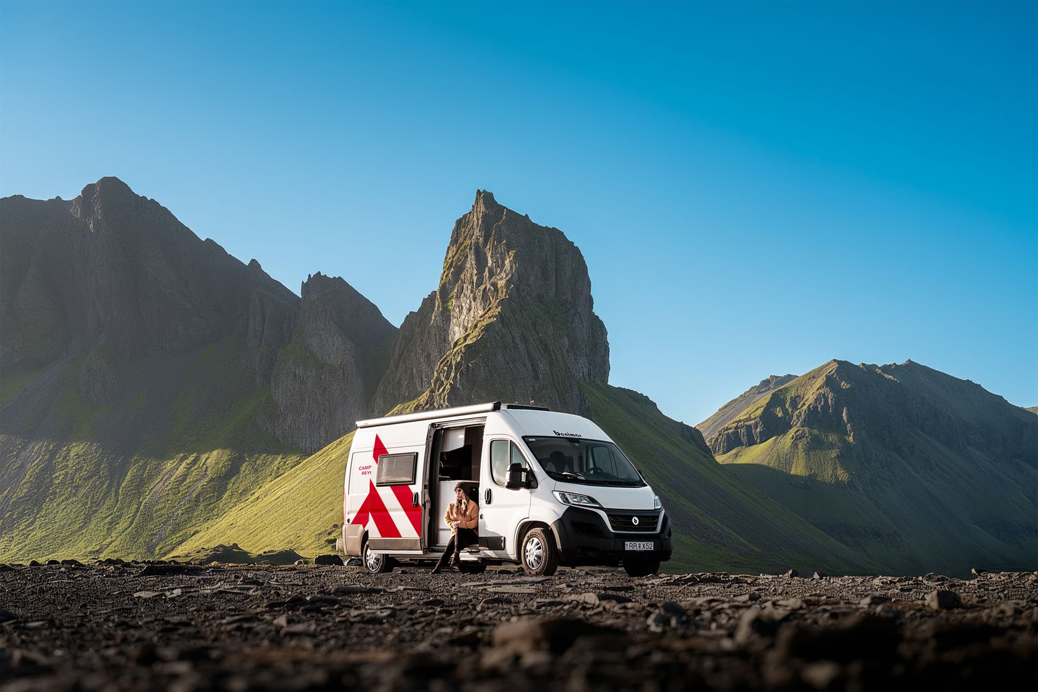 White Campervan Reykjavik rental on black gravel, side door open with traveler seated; green volcanic peaks and clear summer sky in Iceland.