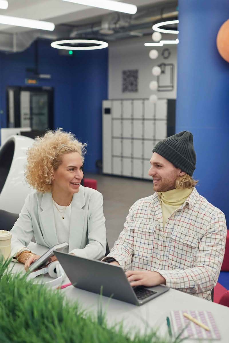 Two friendly support agents at a modern desk, smiling and chatting beside a laptop and headset, illustrating 24/7 multilingual customer support.