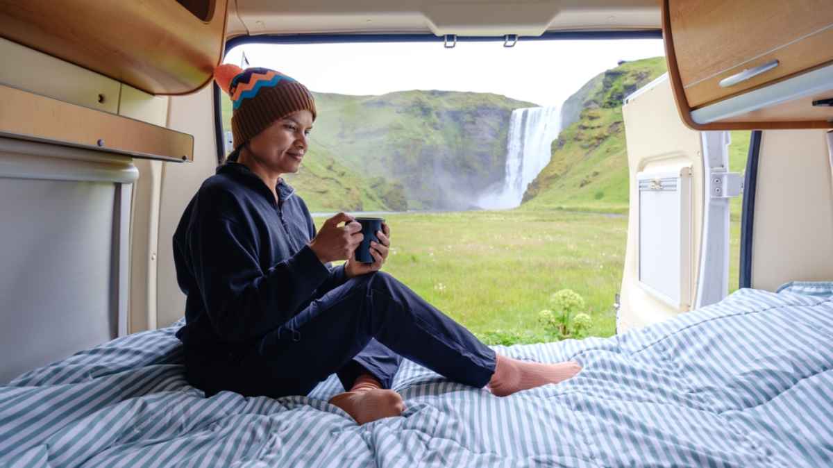 Traveler sipping coffee on a campervan bed with a waterfall view through the back doors.