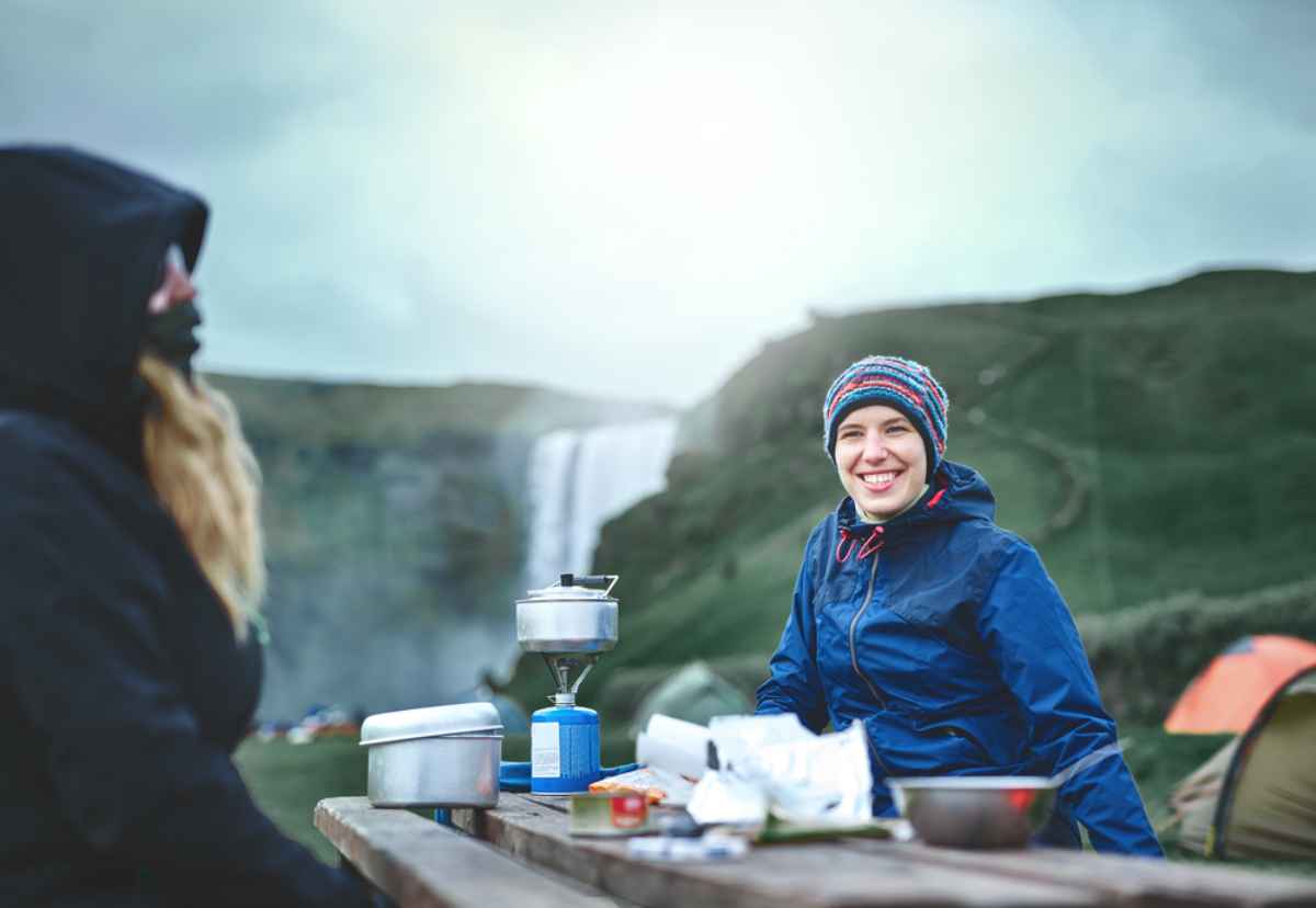 Two people smile near a waterfall, sitting at a picnic table with camping gear. One wears a blue jacket and beanie. Skogafoss in the background.