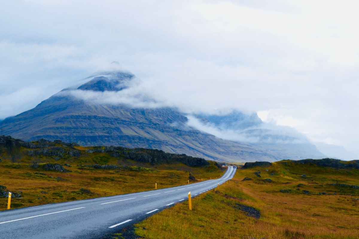 Two-lane Icelandic road curving across mossy plains toward a cloud-shrouded mountain on a cool, misty day.
