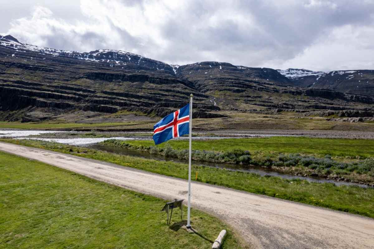 Icelandic flag waving by a gravel road and stream, with layered volcanic mountains and late-spring snow patches under cloudy skies.