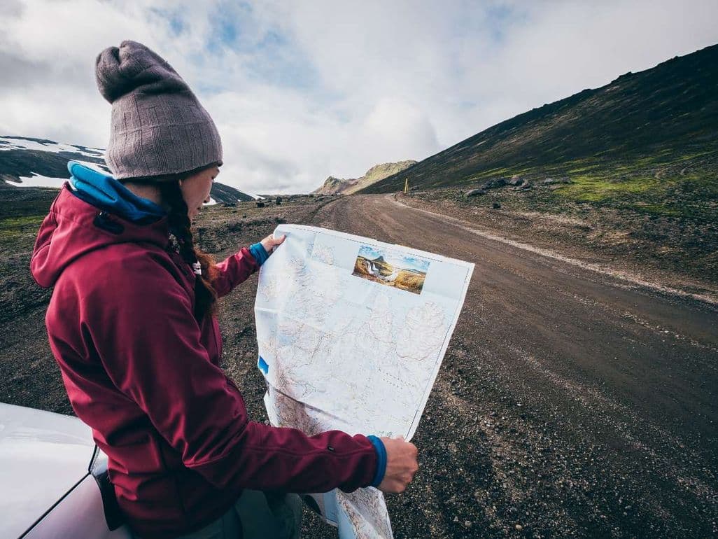 A woman wearing a hat and jacket stands next to a car on a gravel road in Iceland, holding and examining a large map with mountains and green hills in