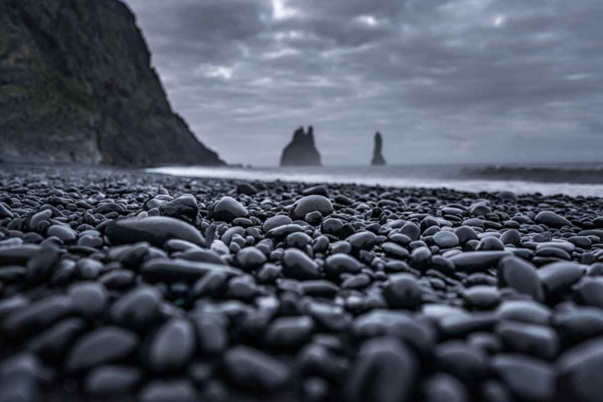 Close-up of wet black pebbles on Reynisfjara beach, Reynisdrangar sea stacks blurred under gray skies.