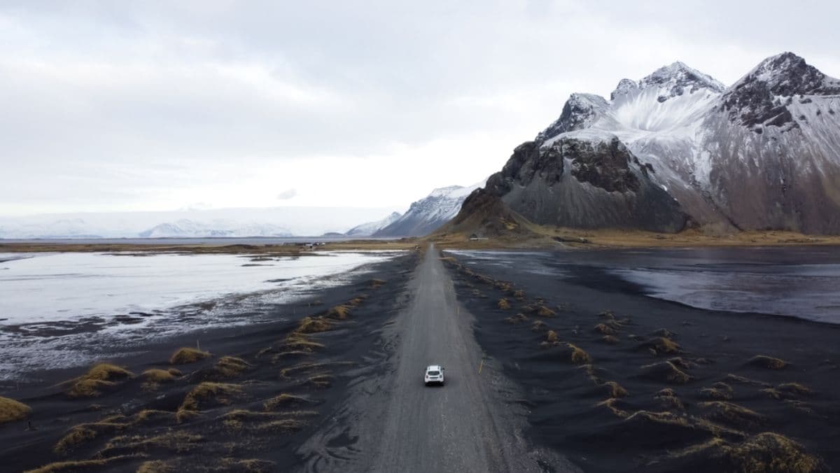 Lonely white car drives a gravel road across black sand dunes toward snow-covered mountains and a frozen lagoon in Iceland under an overcast sky.