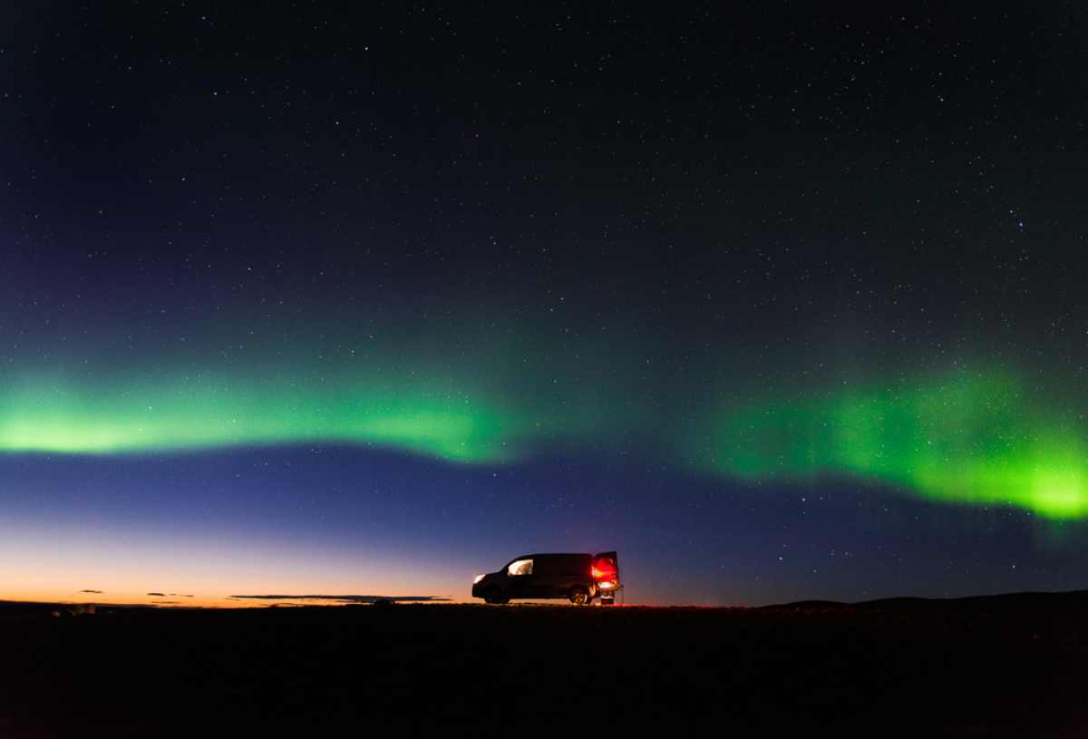 Campervan parked under the green aurora borealis at twilight, starry Icelandic sky above.