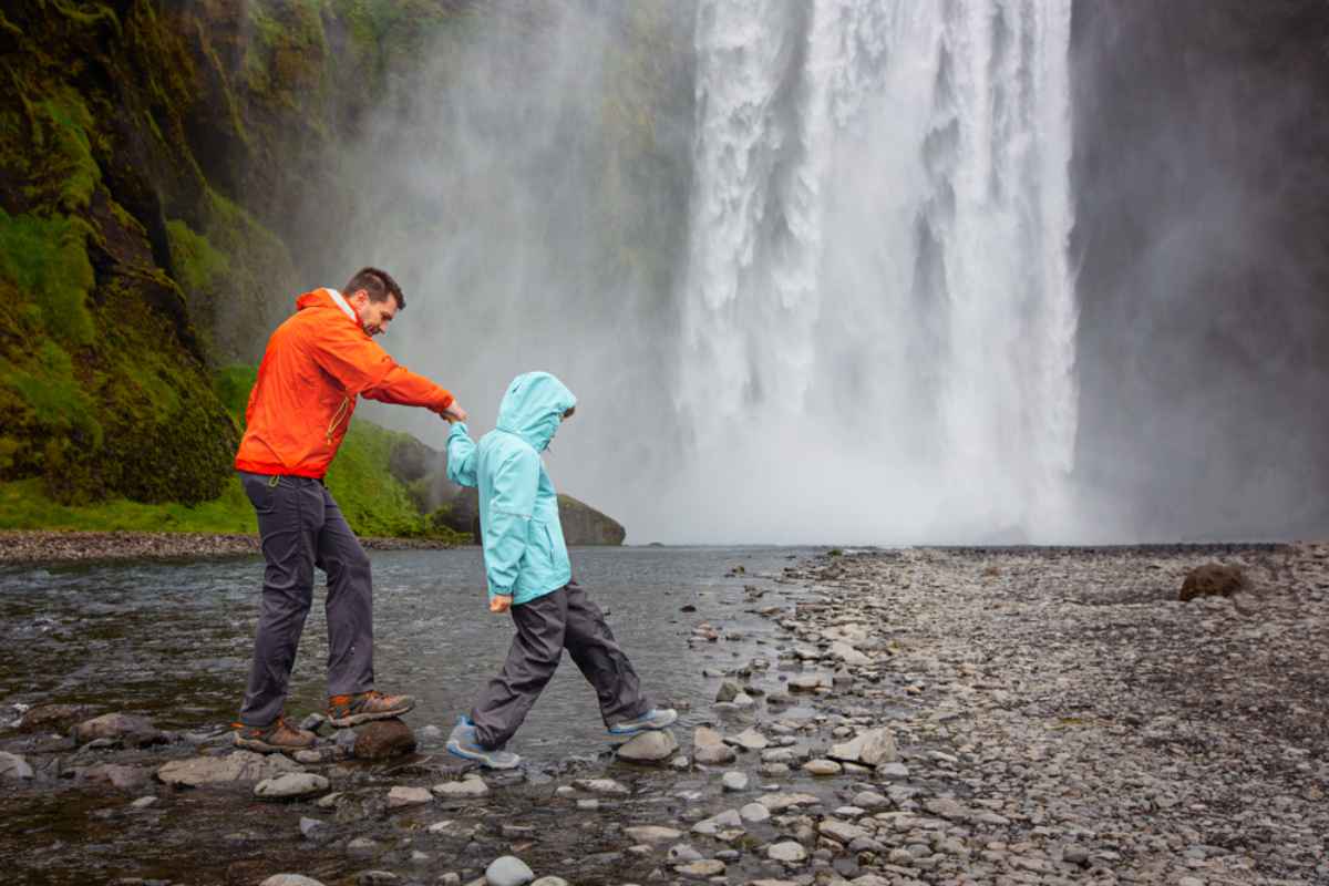 A man in an orange rain jacket helps a child in a blue waterproof jacket step across a shallow rocky stream in front of a large waterfall in Iceland.