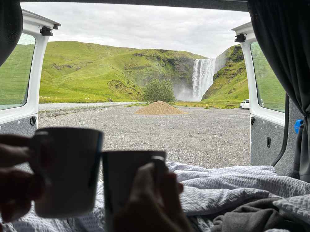 Couple enjoying coffee inside a campervan with a view of a waterfall in Iceland.