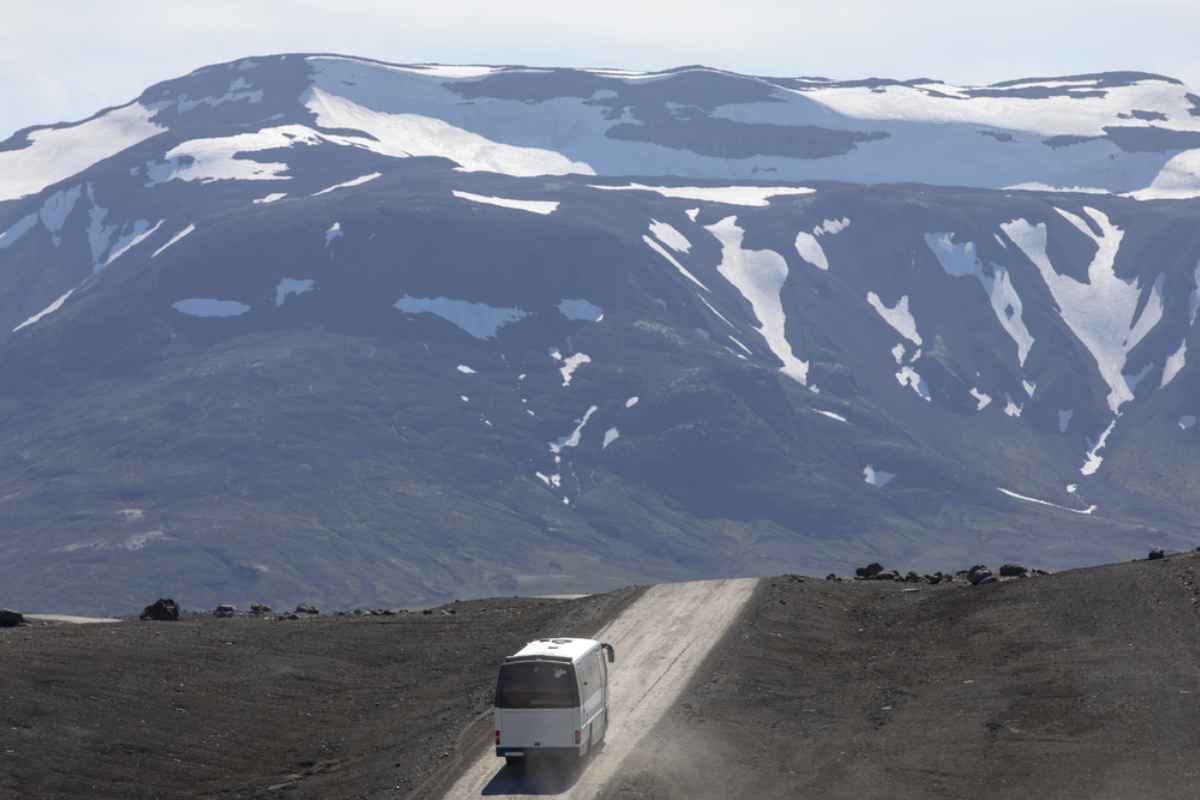 Small bus climbing a remote Icelandic F-road in the Highlands, snow-streaked mountains ahead and dust rising from the gravel.