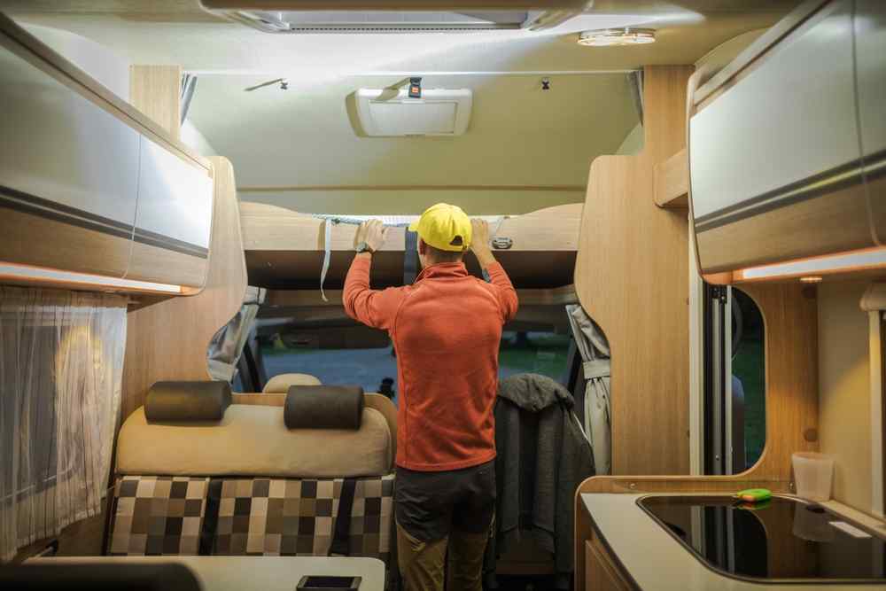Person adjusting an overhead bed inside a modern rental campervan with a seating area, kitchen and storage cabinets.